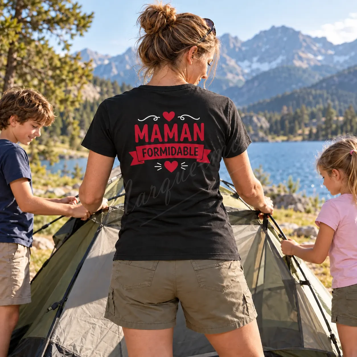 Femme avec des enfants près d’un lac de montagne, portant un t-shirt noir avec le motif « Maman formidable » rouge dans le dos.