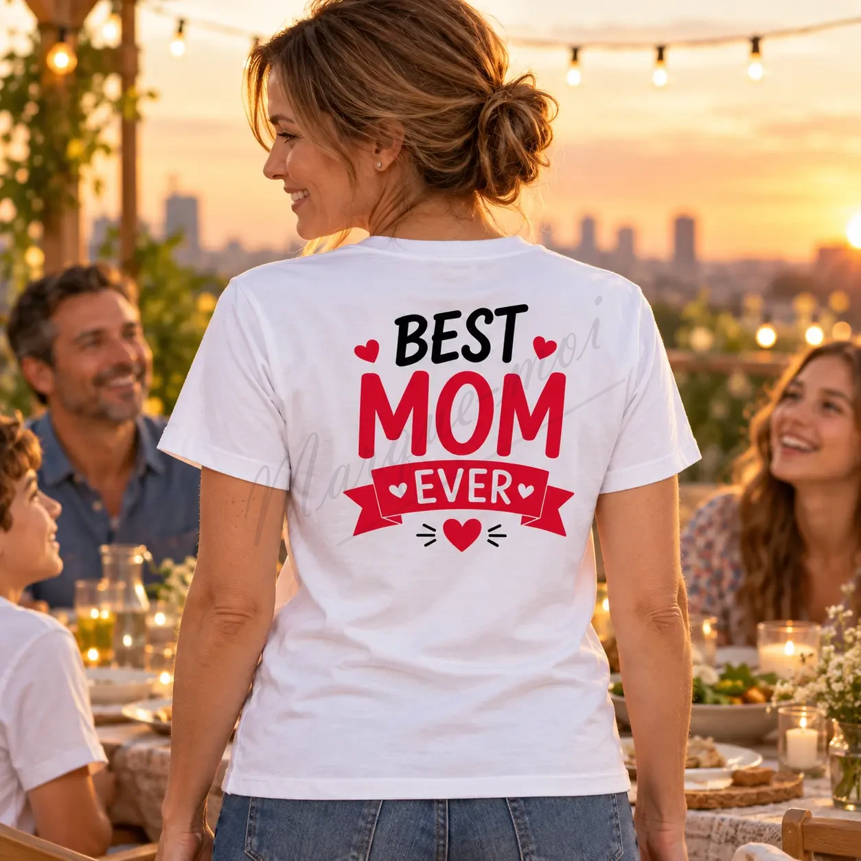 Femme en extérieur sous une guirlande lumineuse, portant un t-shirt blanc avec le motif « Best Mom Ever » noir et rouge dans le dos.