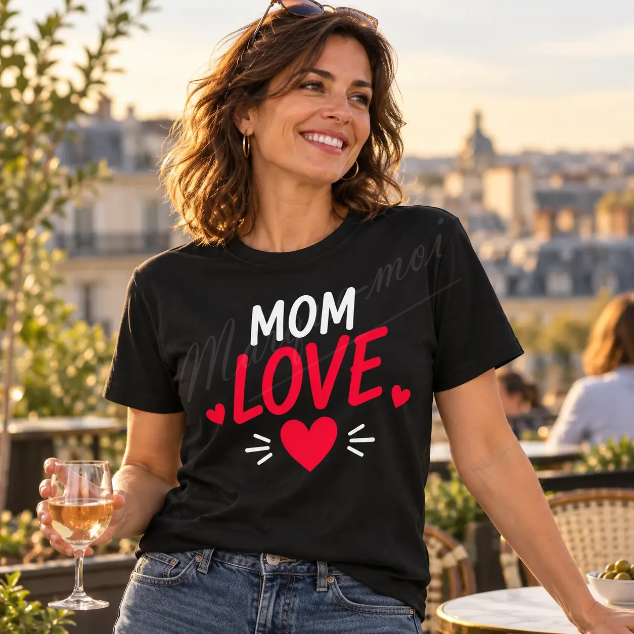 Femme souriante en extérieur, devant un panorama urbain, portant un t-shirt noir « Mom Love » avec un motif blanc et rouge sur le devant.