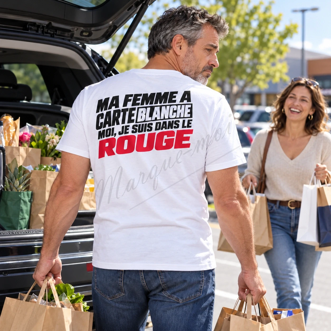 Homme portant un t-shirt blanc avec le slogan « Ma femme a carte blanche, moi je suis dans le rouge » en noir et rouge dans le dos, près d’un coffre de voiture ouvert et d’une femme avec des sacs.