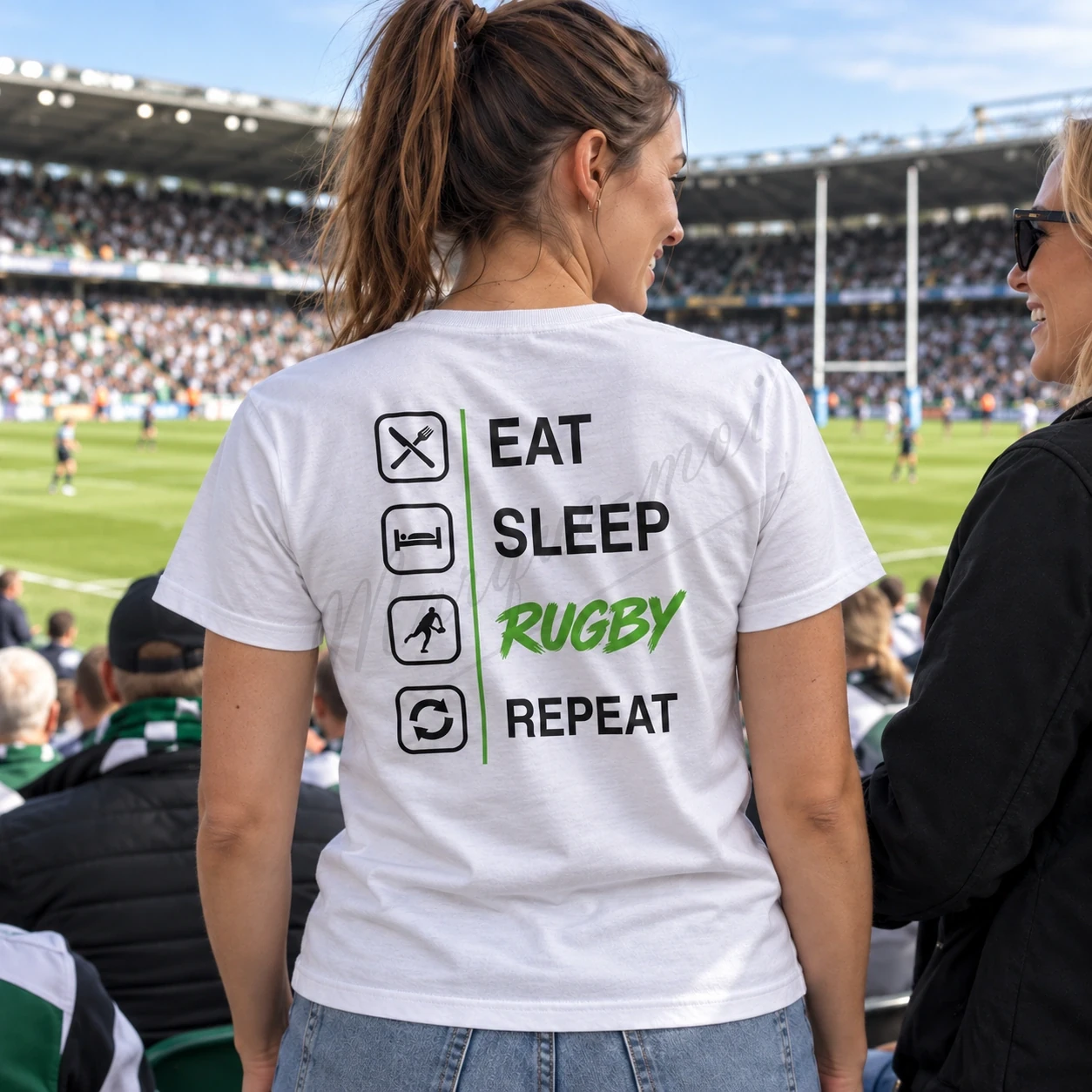 Femme vue de dos dans les tribunes d’un stade de rugby, portant un t-shirt blanc « Eat Sleep Rugby Repeat » au motif noir et vert.