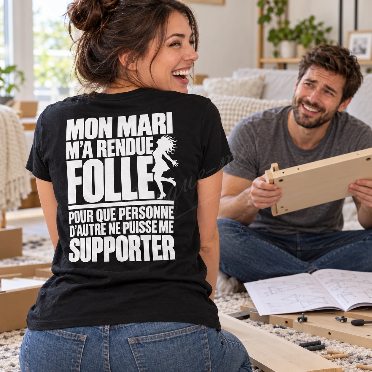 Femme vue de dos dans un salon, en t-shirt noir « Mon mari m’a rendue folle pour que personne d’autre ne puisse me supporter », face à un homme assis.
