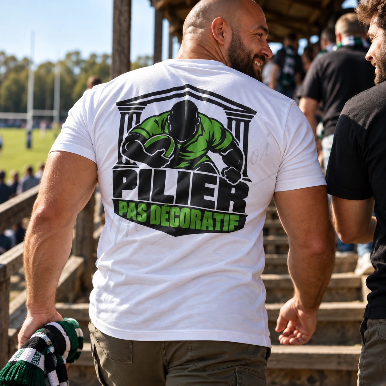 Homme vu de dos près des tribunes d’un terrain de rugby, portant un t-shirt blanc « Pilier, pas décoratif » au motif noir et vert.