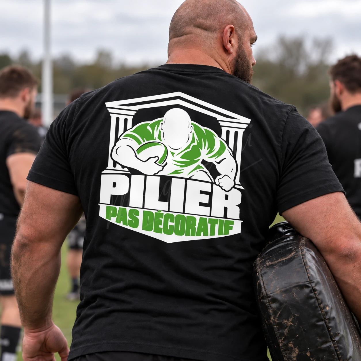 Homme vu de dos près d’un terrain de rugby, portant un t-shirt noir « Pilier, pas décoratif » au motif blanc et vert clair.