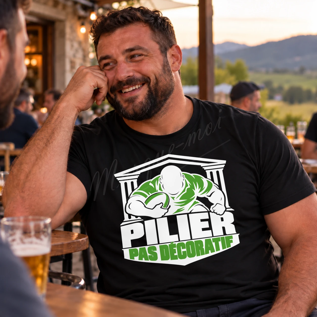 Homme assis en terrasse portant un t-shirt noir « Pilier, pas décoratif », avec un joueur de rugby illustré en blanc et vert.